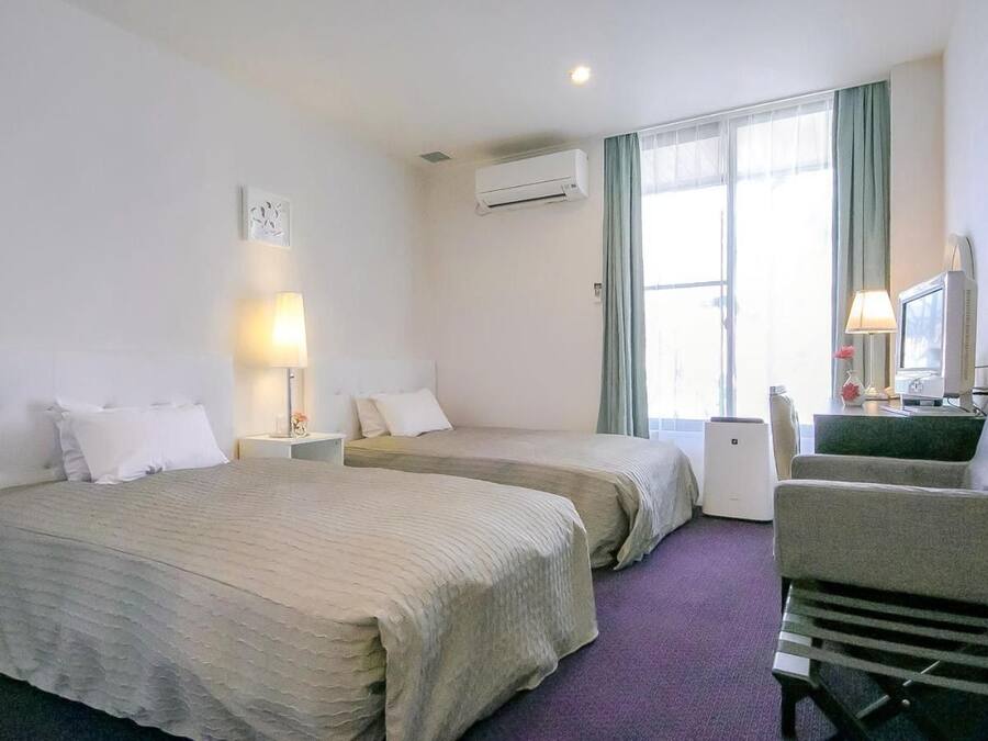 Una habitación de hotel con dos camas, un escritorio con una computadora, una silla y un ventanal con cortinas.