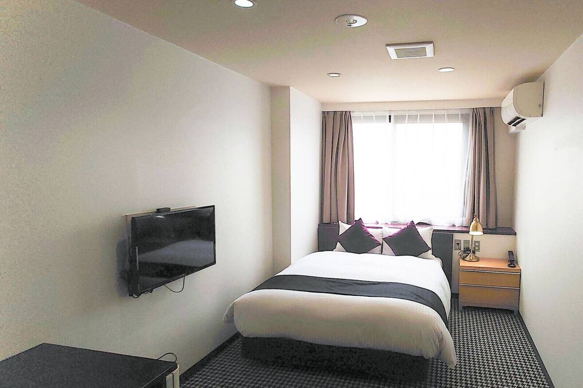 A hotel room with a bed, a TV mounted on the wall, a desk with a lamp, and a window with curtains.