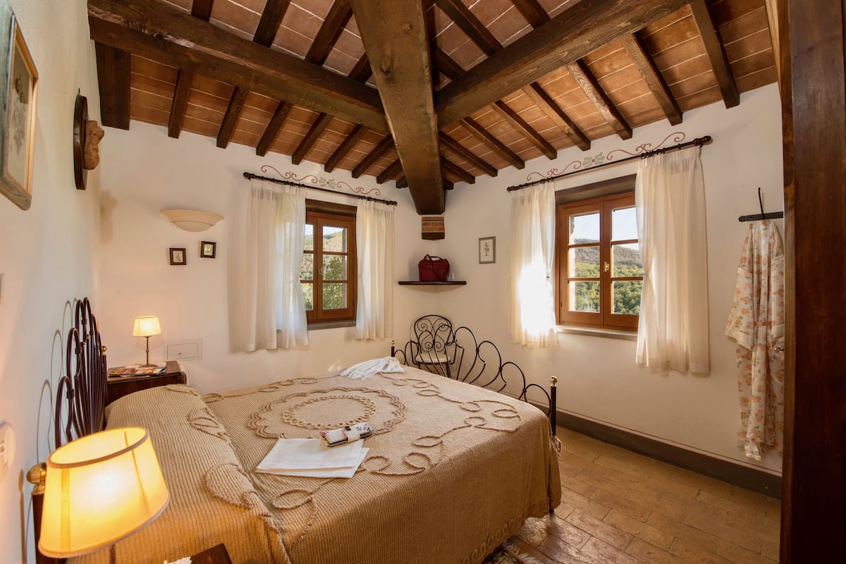 Un dormitorio con techo de madera, una cama con colcha estampada, dos ventanas con cortinas y una mesita de noche con lámpara.