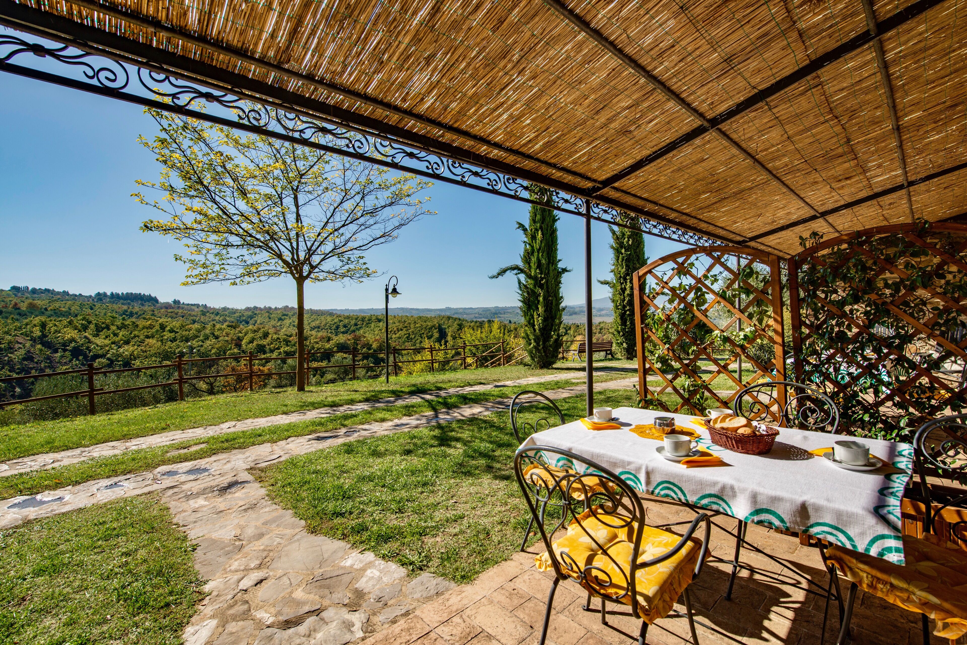 Un espace de restauration en plein air avec une table dressée pour deux personnes, entourée de chaises, sous une pergola offrant une vue sur un paysage luxuriant.