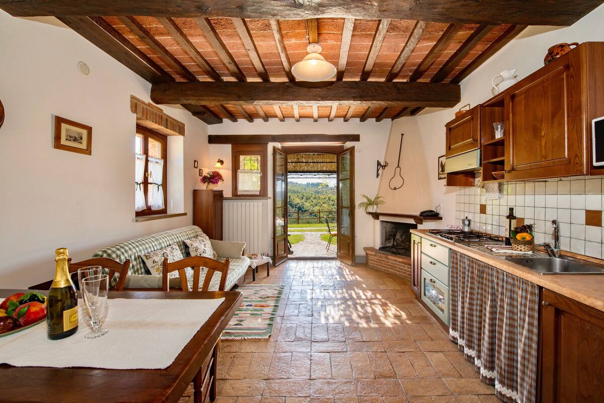 Una cocina rústica con vigas de madera, un comedor con una mesa dispuesta para dos personas y una vista a través de la puerta abierta.