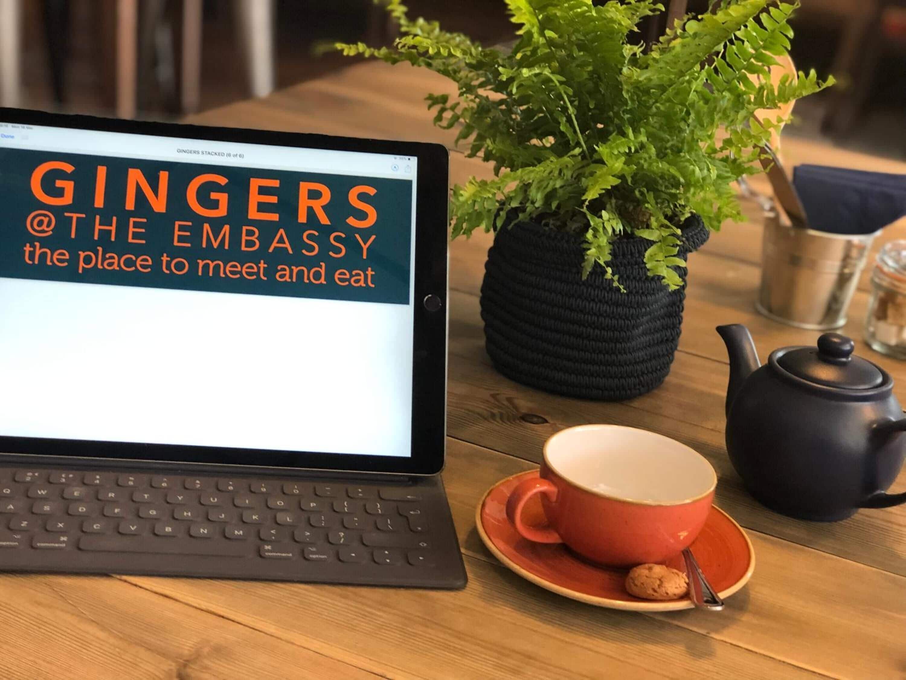 Un ordinateur portable affiche le texte « GINGERS @THE EMBASSY, l’endroit idéal pour se rencontrer et prendre un repas », avec une plante en pot, une théière et une tasse sur une table en bois.