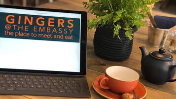 Un ordinateur portable affiche le texte « GINGERS @THE EMBASSY, l’endroit idéal pour se rencontrer et prendre un repas », avec une plante en pot, une théière et une tasse sur une table en bois.