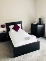 A neatly made bed with white linens and a single purple pillow, a black nightstand with a lamp, and a black dresser with a flat-screen TV.