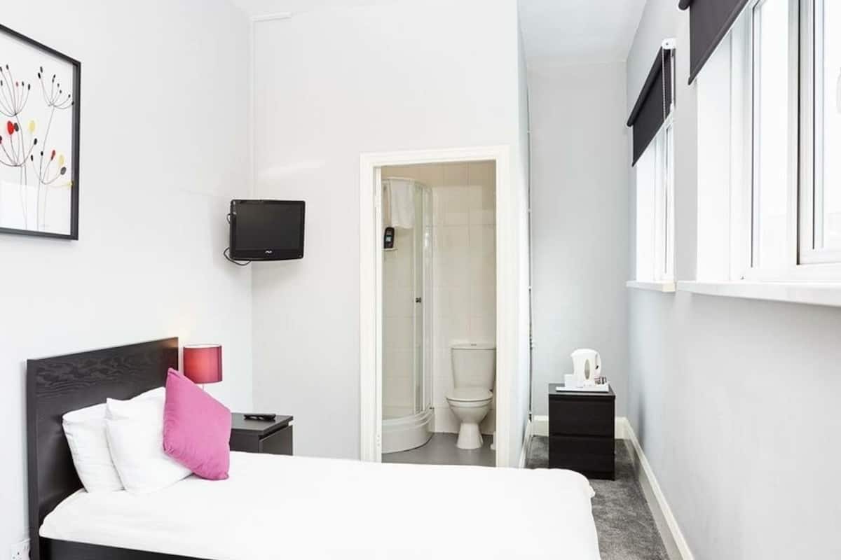 A modern bedroom with a black bed, white walls, a small bedside table with a lamp, a TV mounted on the wall, and a view of the bathroom through an open door.