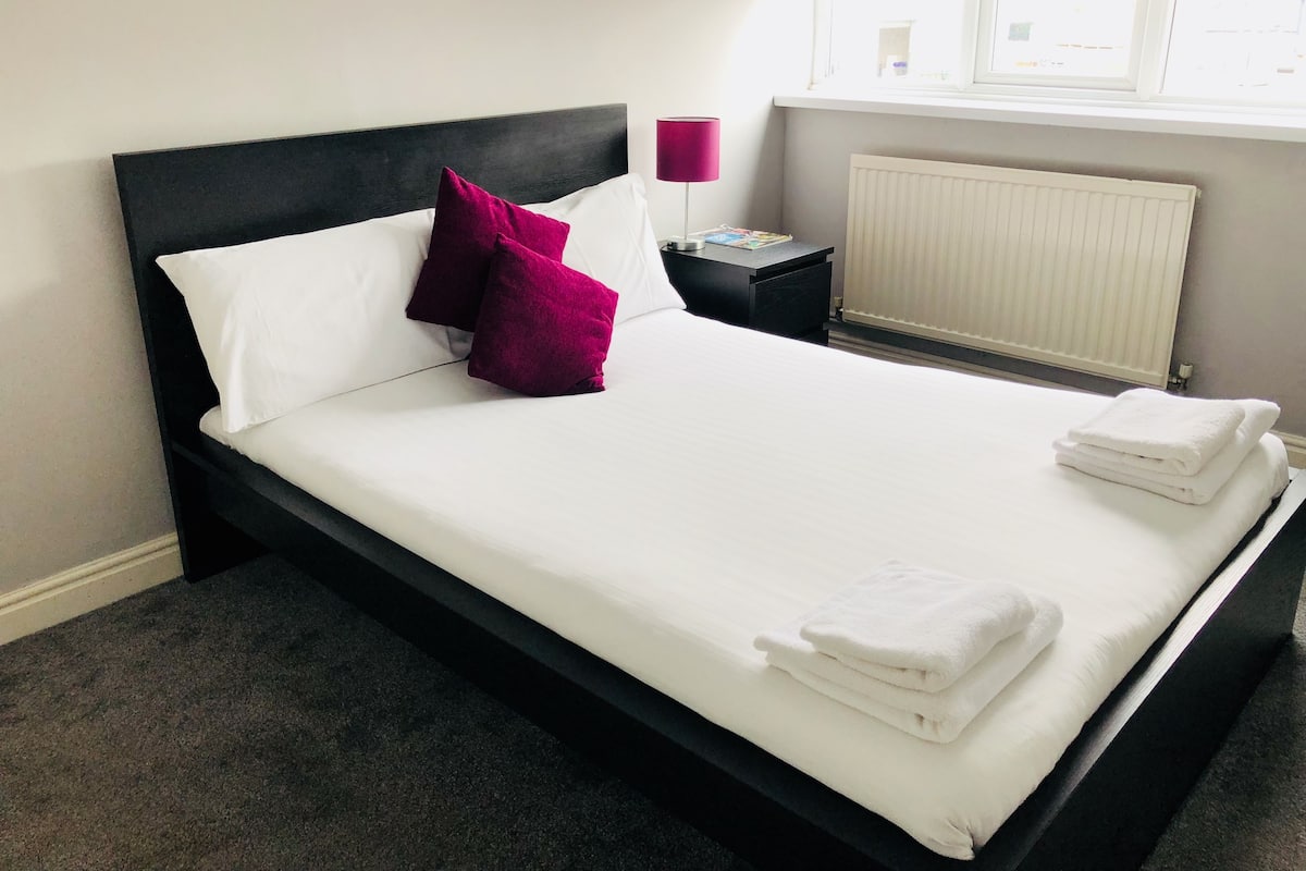 A neatly made bed with white linens and two pillows, a bedside table with a lamp, and a window with blinds.