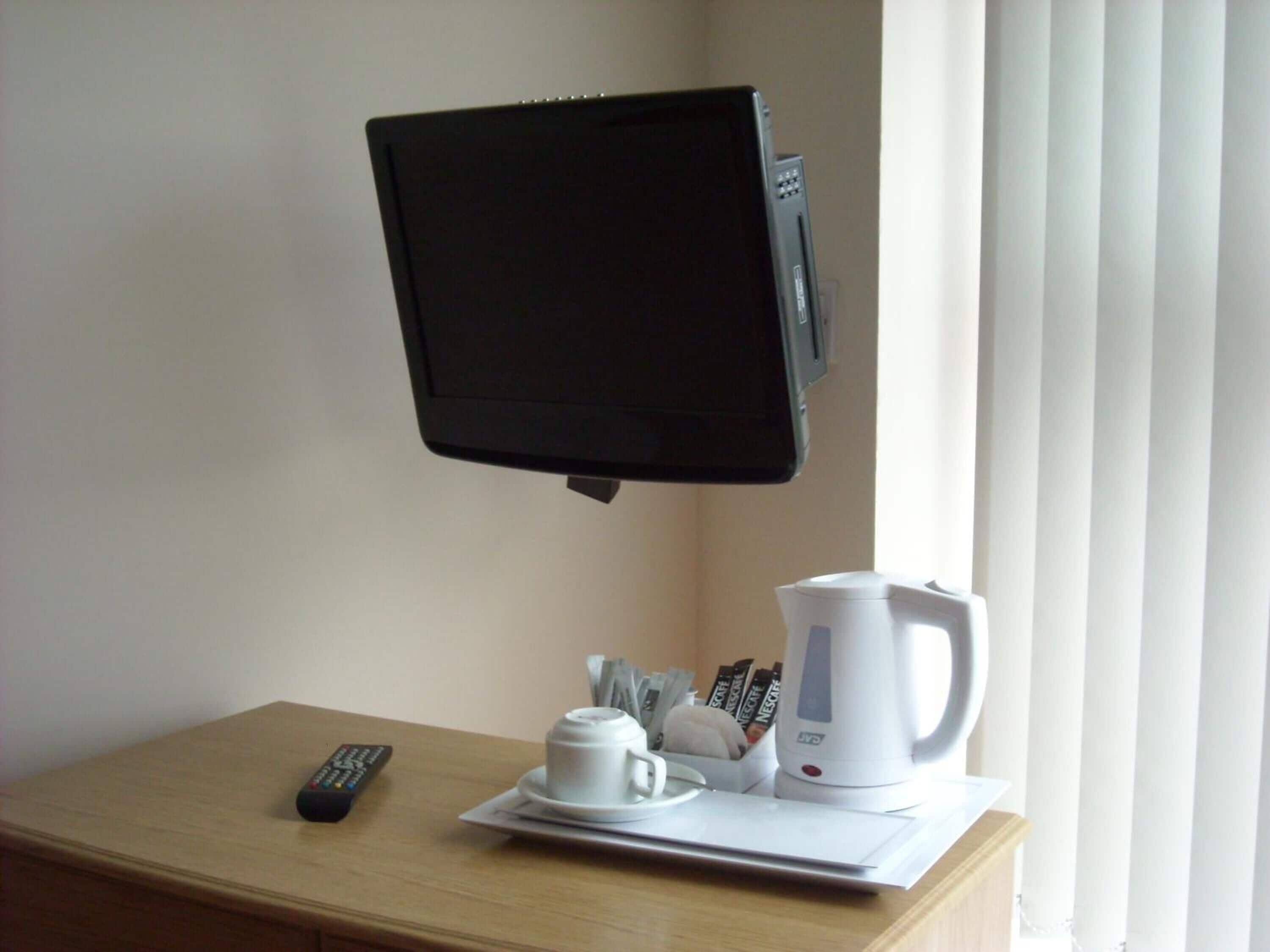 A room with a mounted television, a remote control, a white coffee maker, and a tray with a cup and saucer on a wooden table.