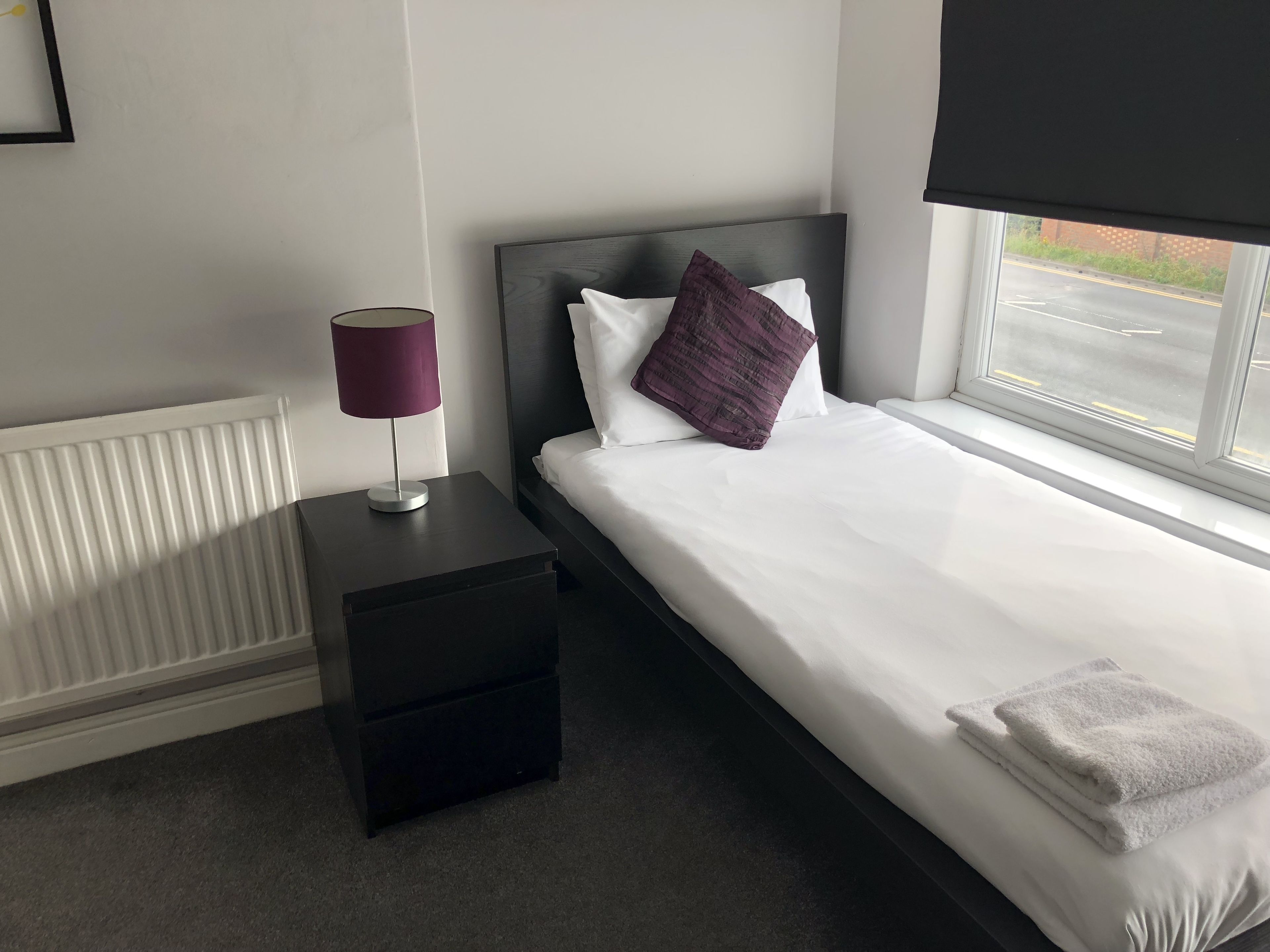 A neatly made bed with white linens and a folded white towel, a dark nightstand with a lamp, and a window with a view of the street.