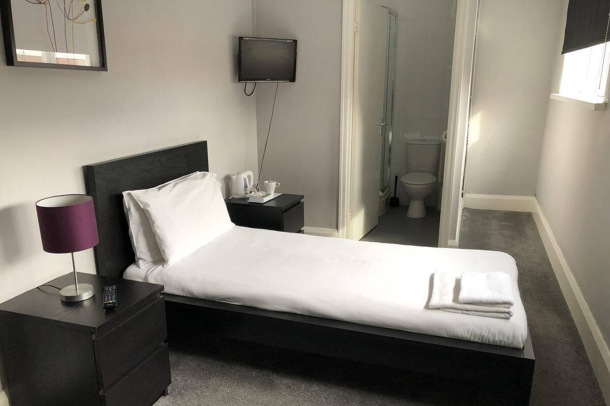 A neatly made bed with white linens, a black headboard, and a folded towel on the footboard. There is a small bedside table with a lamp and a remote control. A television is mounted on the wall above the bed.