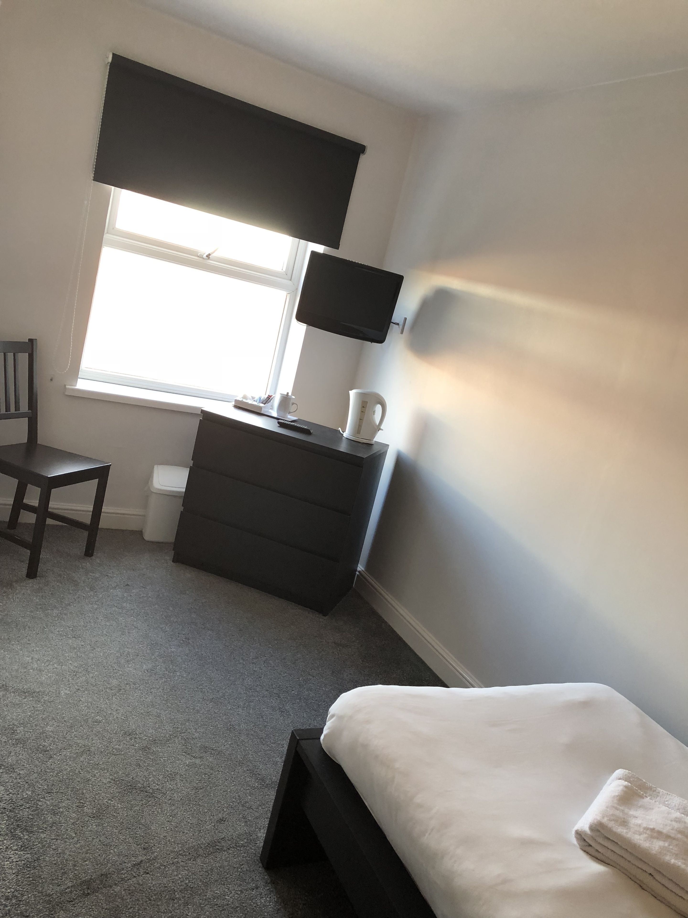 A bedroom with a bed, a desk with a chair, a window with blinds, and a television on the wall.