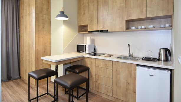 Uma cozinha compacta com armários de madeira, bancada branca, pia, micro-ondas, chaleira e um pequeno frigorífico. Há duas banquetas e uma luminária pendente.