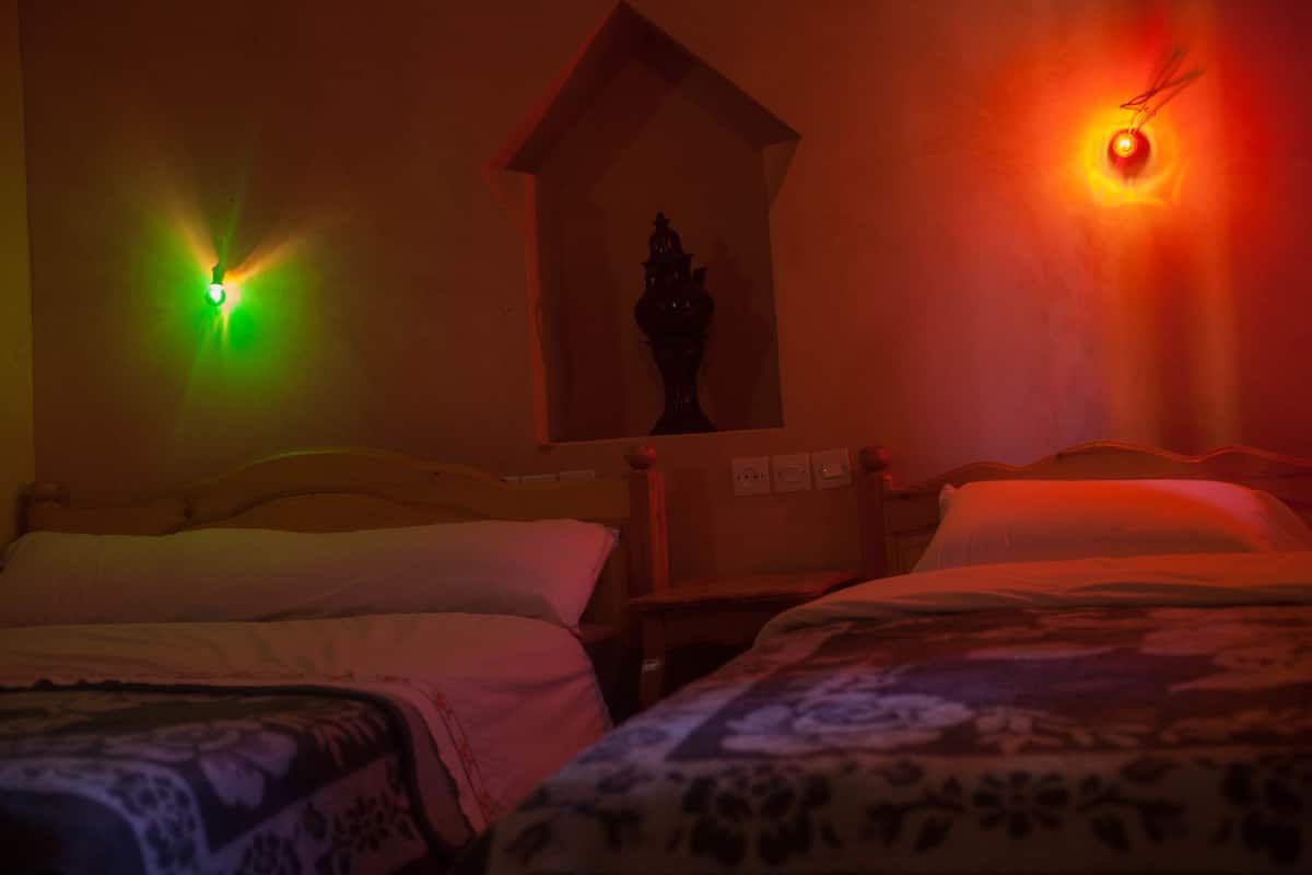 A dimly lit room with two beds, a small table, and a wall-mounted shelf with a decorative item.