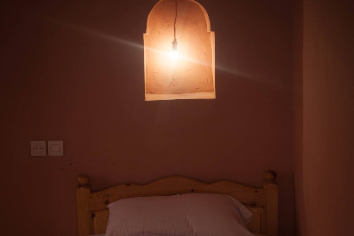 A dimly lit room with a single bed, a hanging light fixture, and a power outlet on the wall.