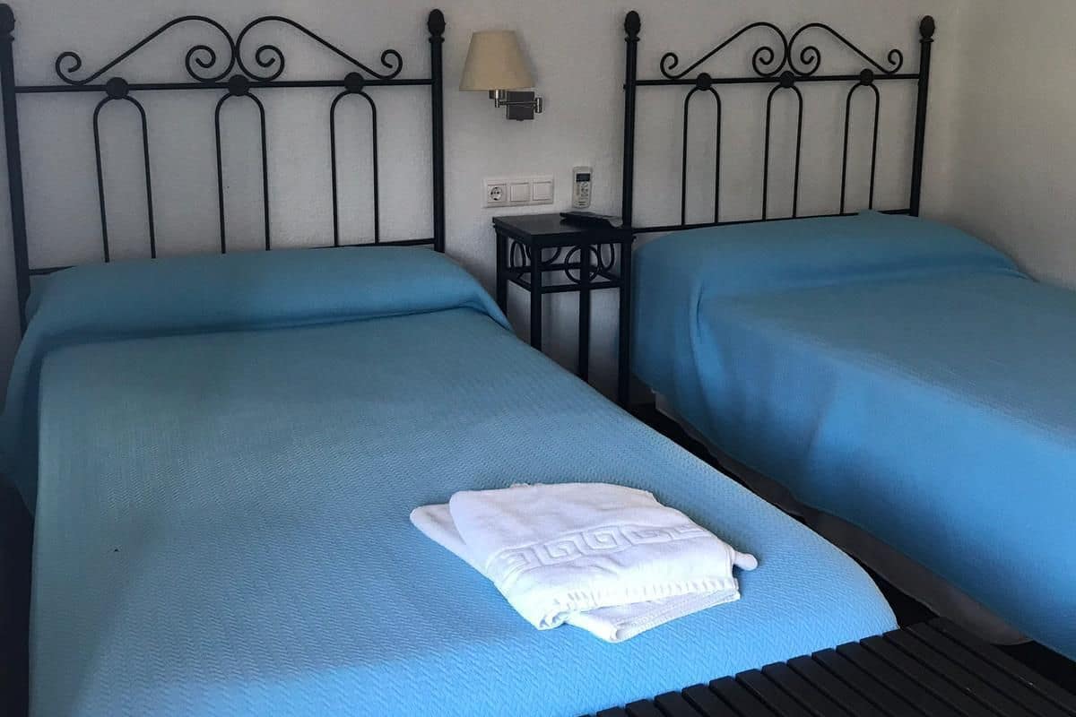 A room with two beds, each with a blue bedspread and a white towel on the bed in the middle. There is a small table with a lamp between the beds.