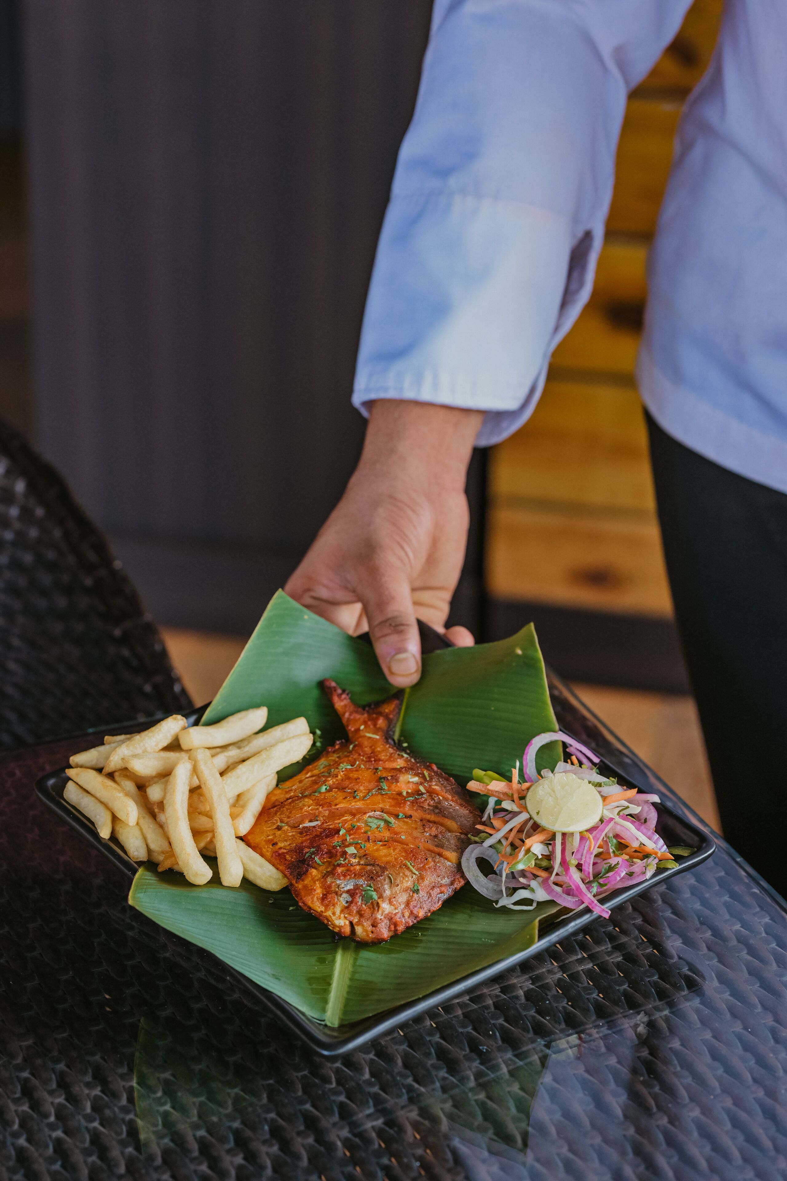 A person holding a tray with a grilled fish, fries, and a salad on a black textured table.