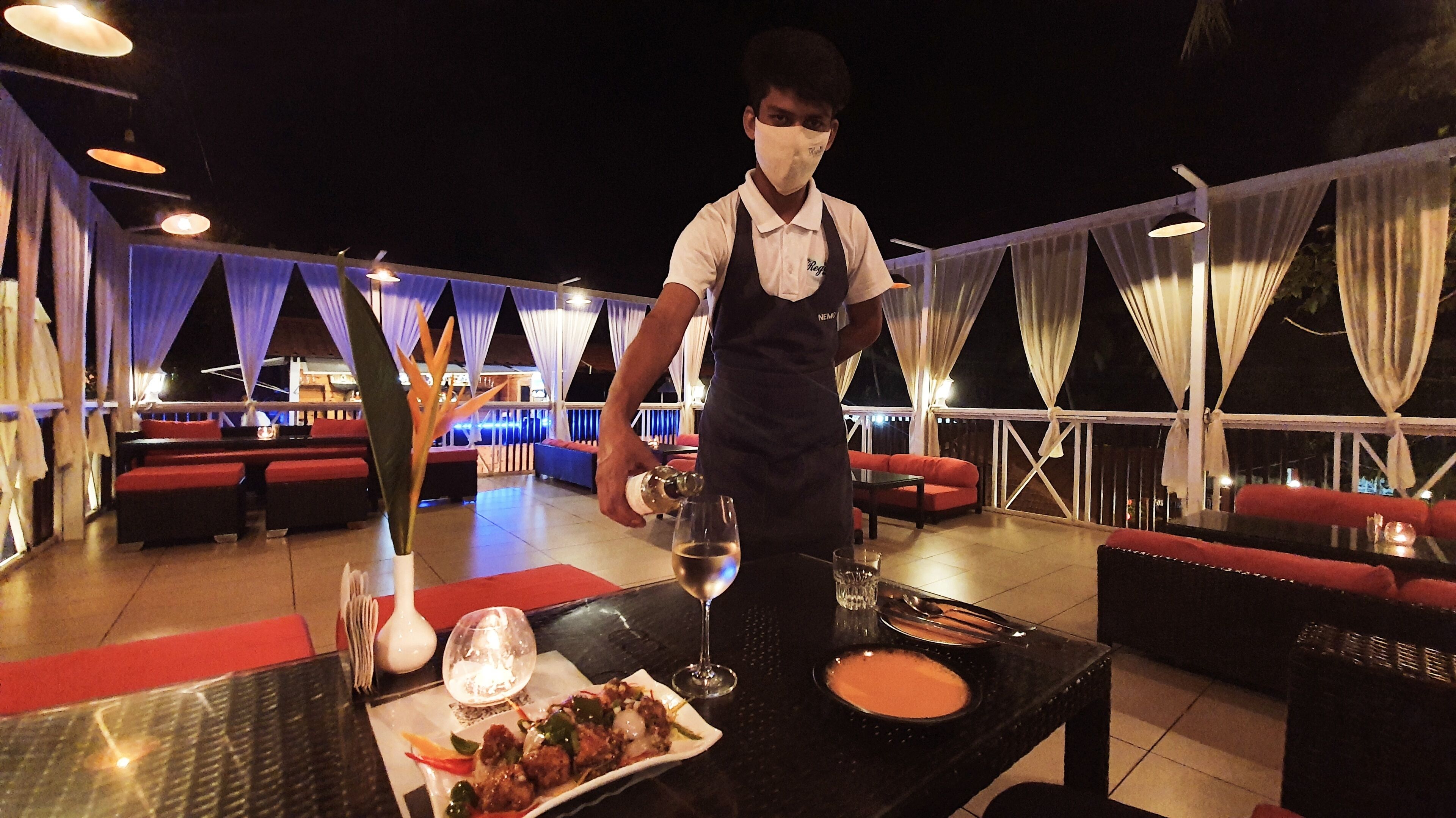 A restaurant with outdoor seating, a waiter pouring a drink, and a table set with a plate of food and a glass of wine.