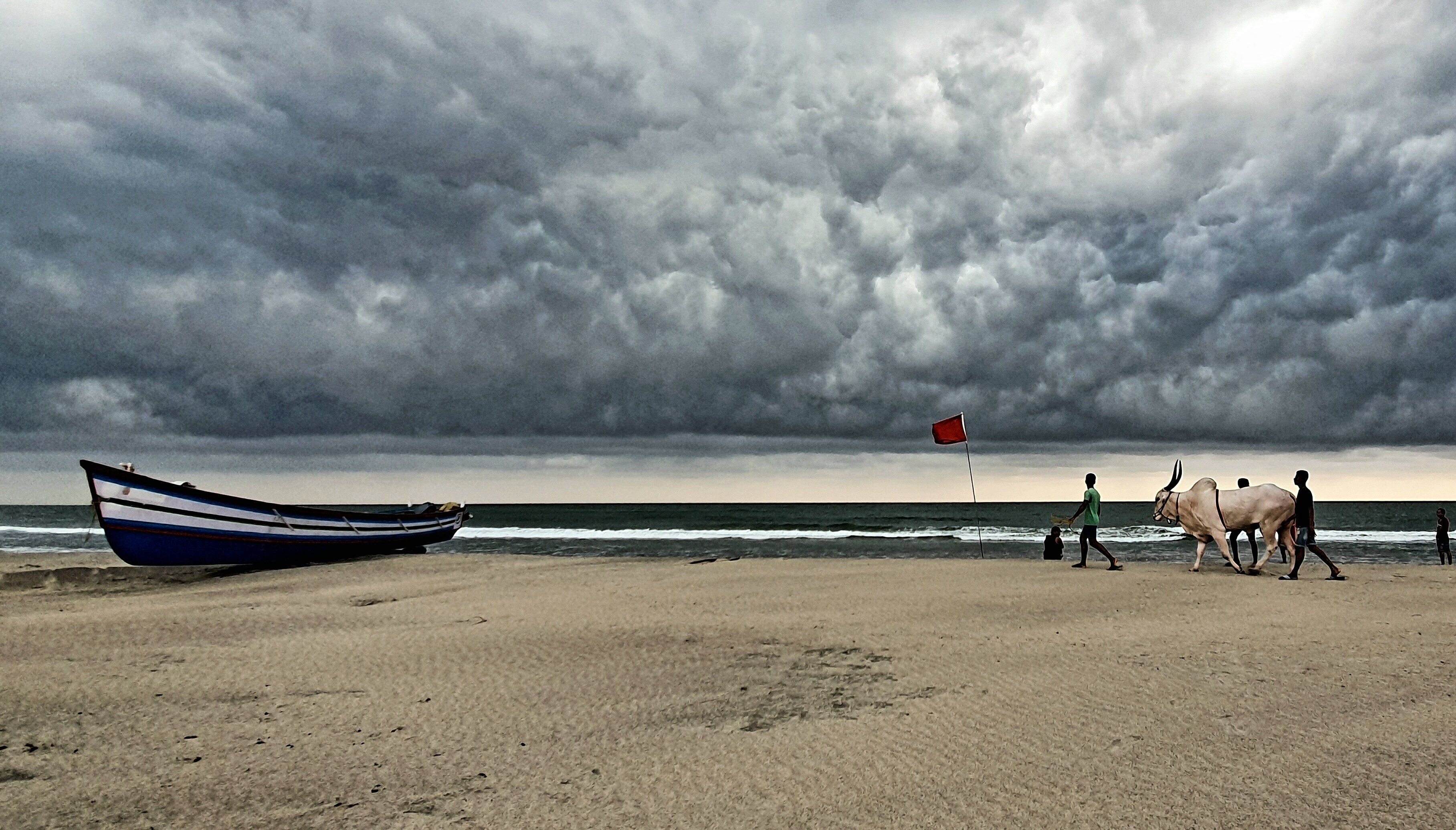 Una playa con un bote, una bandera roja y gente caminando.
