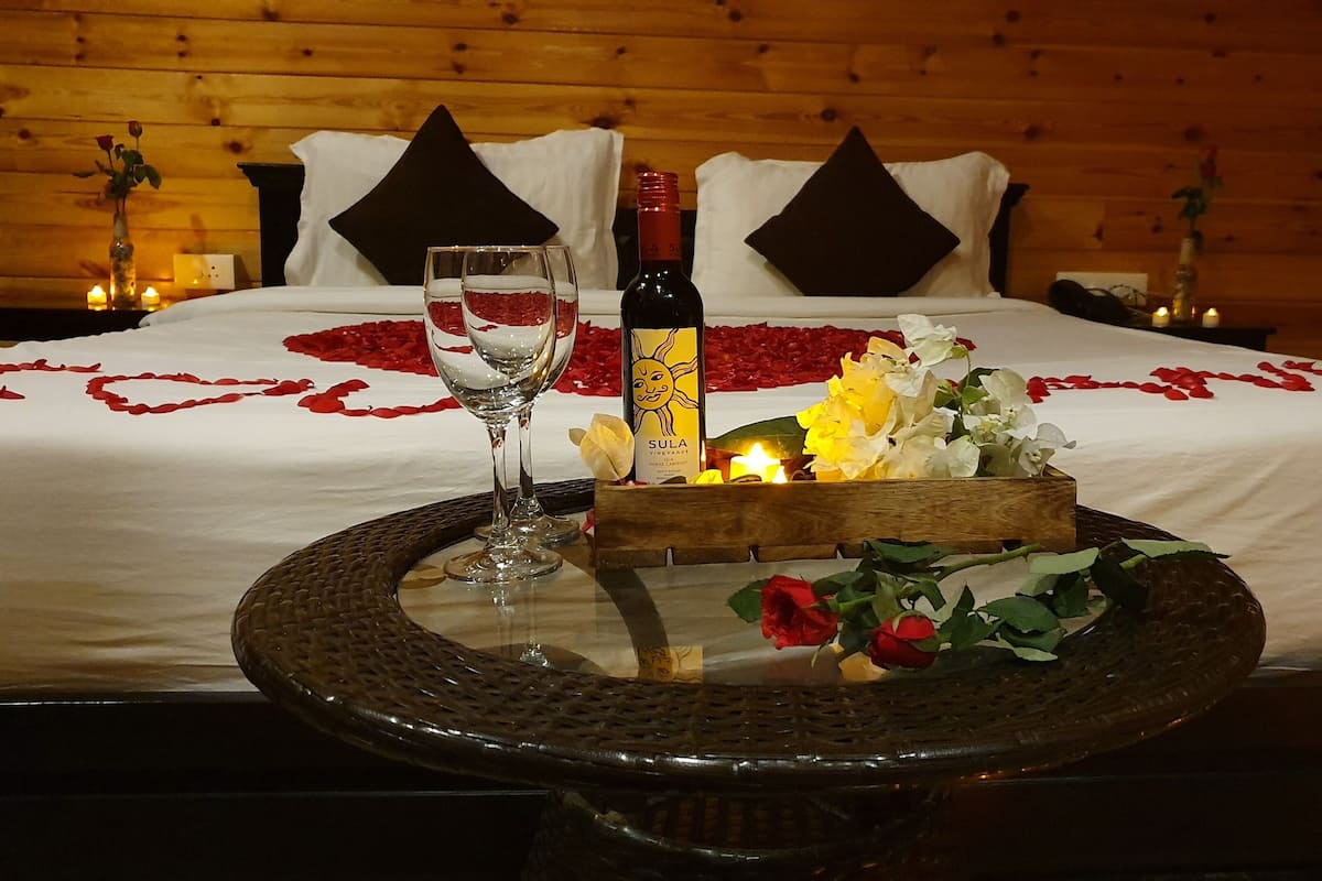 A neatly made bed with a white bedspread, red rose petals, a bottle of Sula Vineyards wine, and a glass of water on a round table.