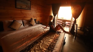 A wooden cabin room with a bed, a painting on the wall, and a view of the outdoors through a window. - The Regalia Resort (Ashvem)