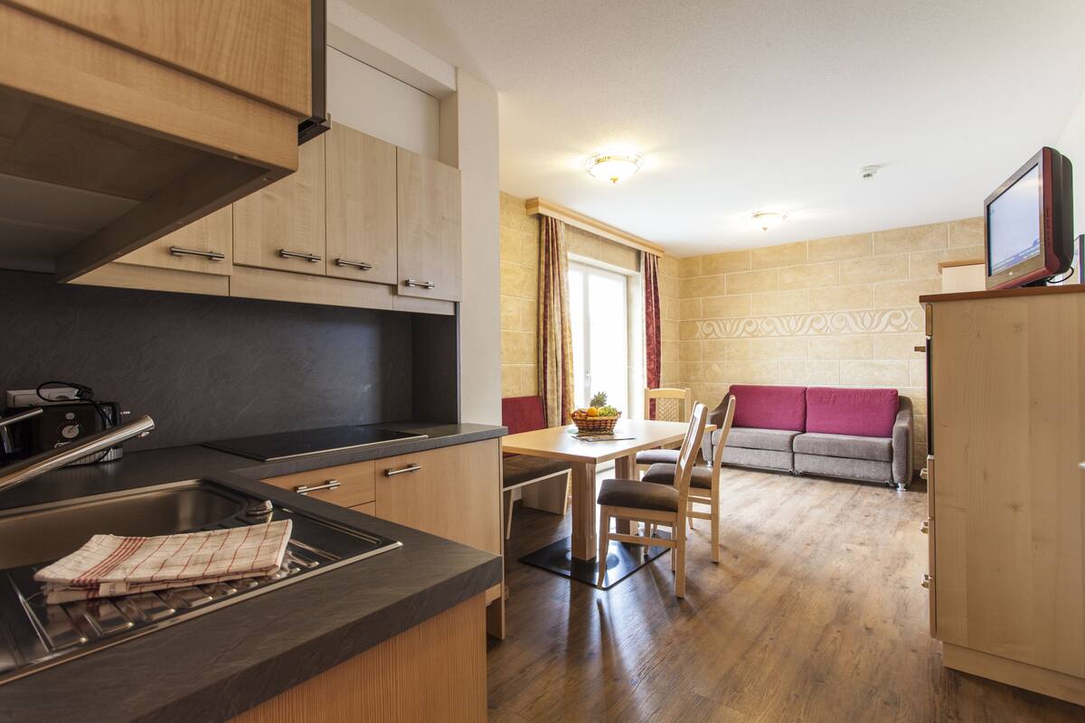 A compact kitchen with a sink, stove, and wooden cabinets. There is a dining area with a table and chairs, and a sofa in the background.