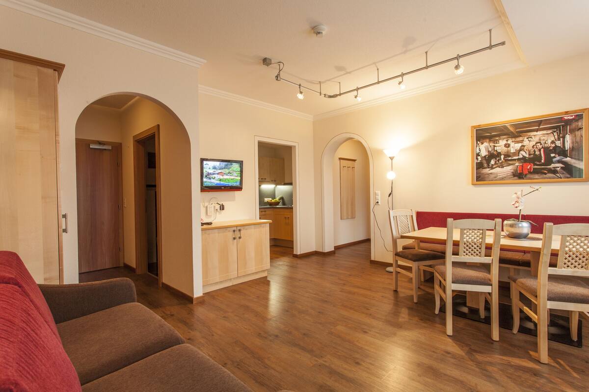 A living room with a red couch, wooden dining table with chairs, a TV, and a kitchen area with cabinets and a countertop.