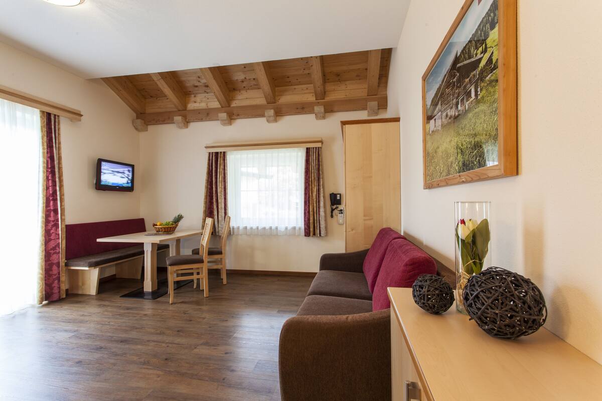 A cozy living room with a wooden ceiling, a sofa, a bench, a dining table with chairs, and a television on the wall.