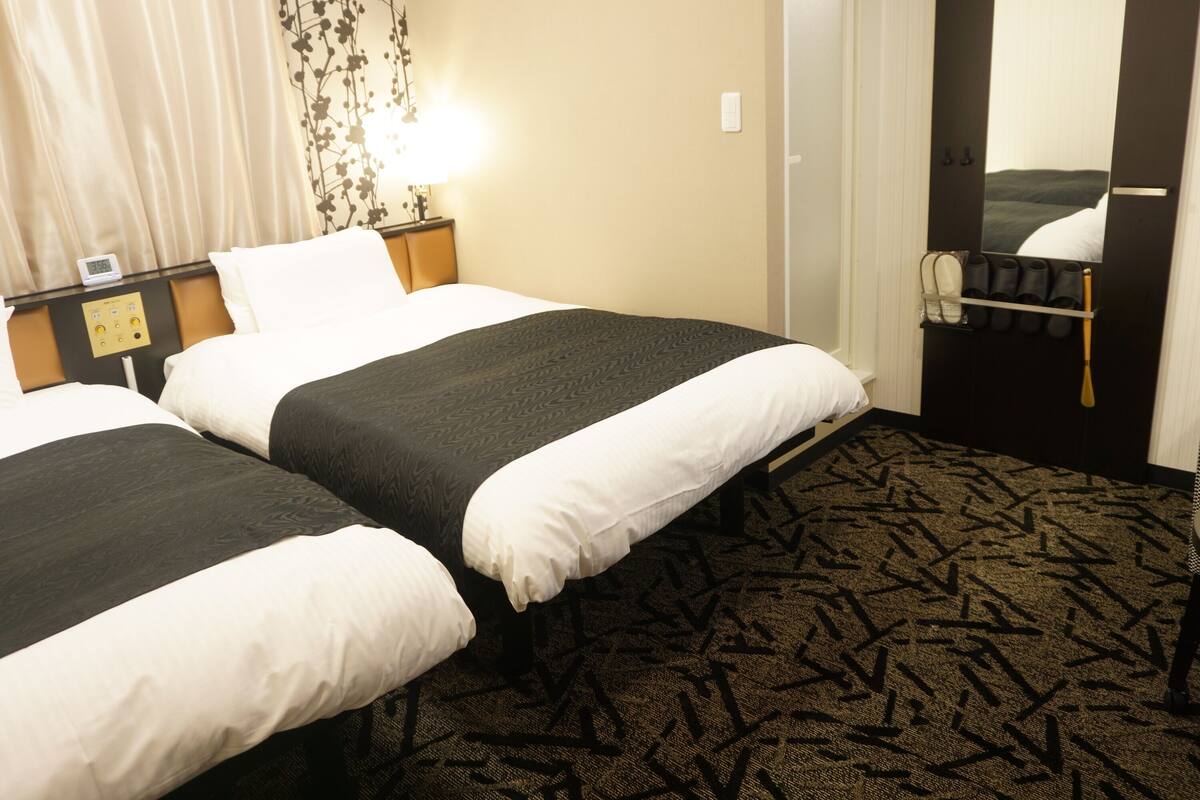 A hotel room with two beds, a nightstand, a lamp, a mirror, and a door with a towel rack.