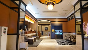 A hotel lobby with a reception desk, a chandelier, a waiting area with a sofa, and a tiled floor.