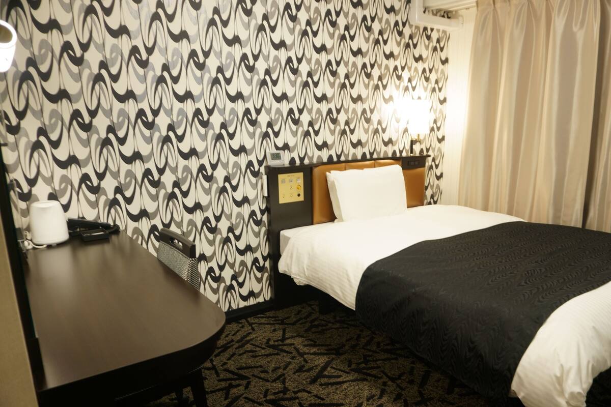 A hotel room with a patterned wallpaper, a bed with white and black bedding, a desk with a telephone and a cup, and a carpet with a geometric design.