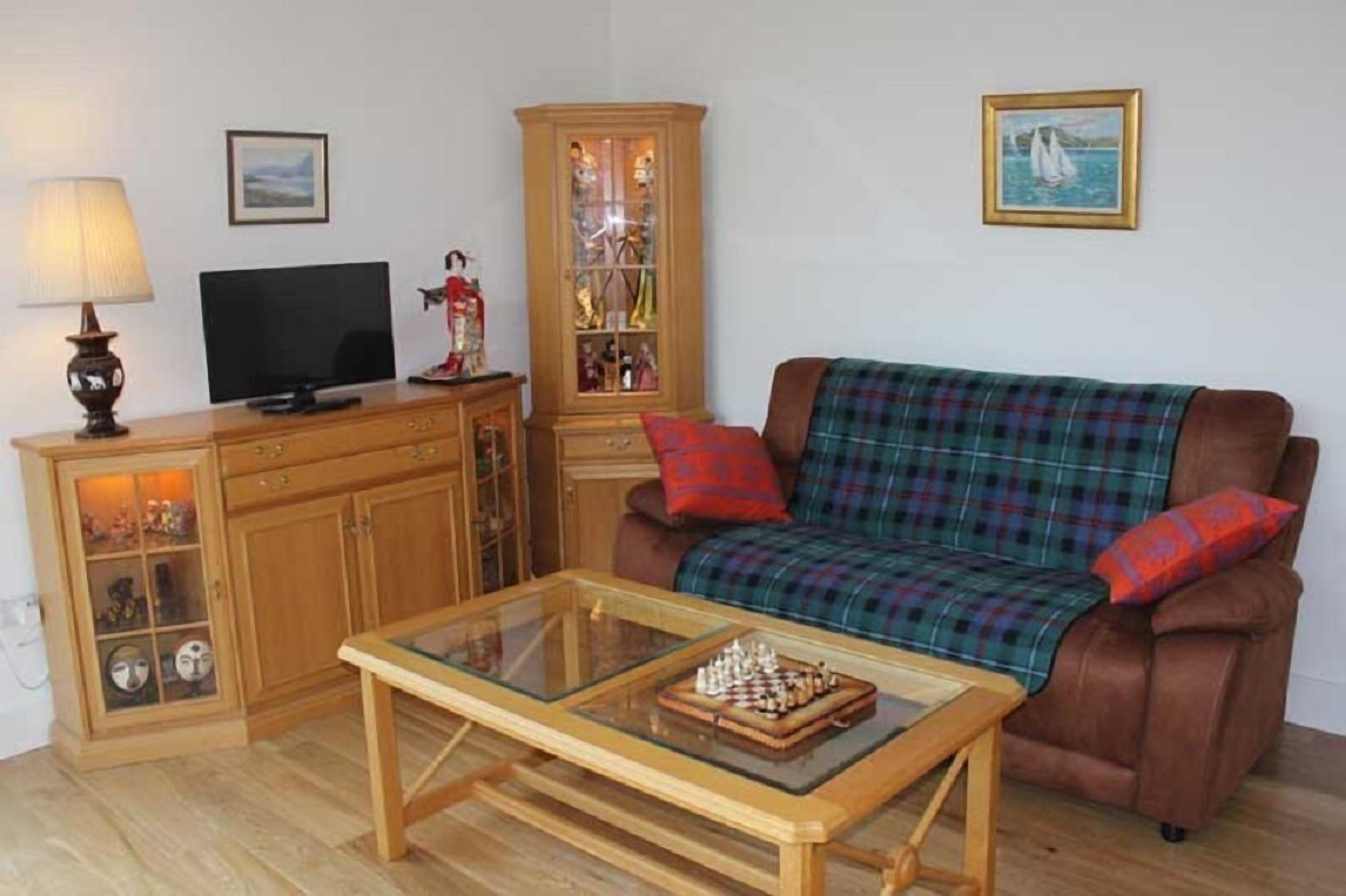A living room with a brown sofa, a glass coffee table with chess pieces, a wooden cabinet with glass doors, a television, and a lamp.