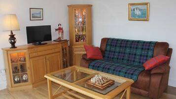 A living room with a brown sofa, a glass coffee table with chess pieces, a wooden cabinet with glass doors, a television, and a lamp.