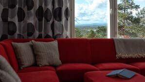 Ein rotes Sofa mit einem Buch, einem gemusterten Vorhang und Blick auf einen Wald und Berge durch ein Fenster.
