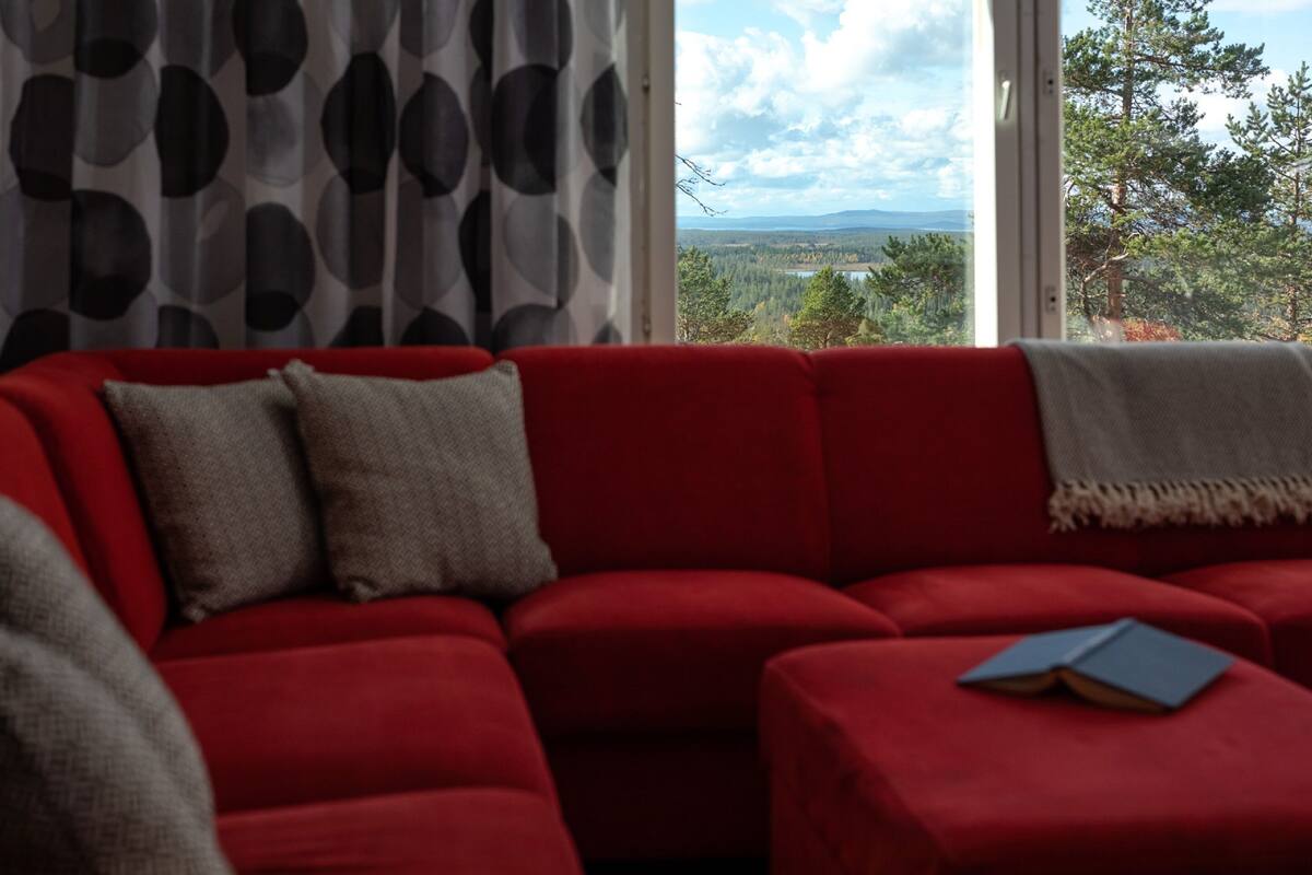 En rød sofa med en bog, et mønstret gardin og udsigt til skov og bjerge gennem et vindue.