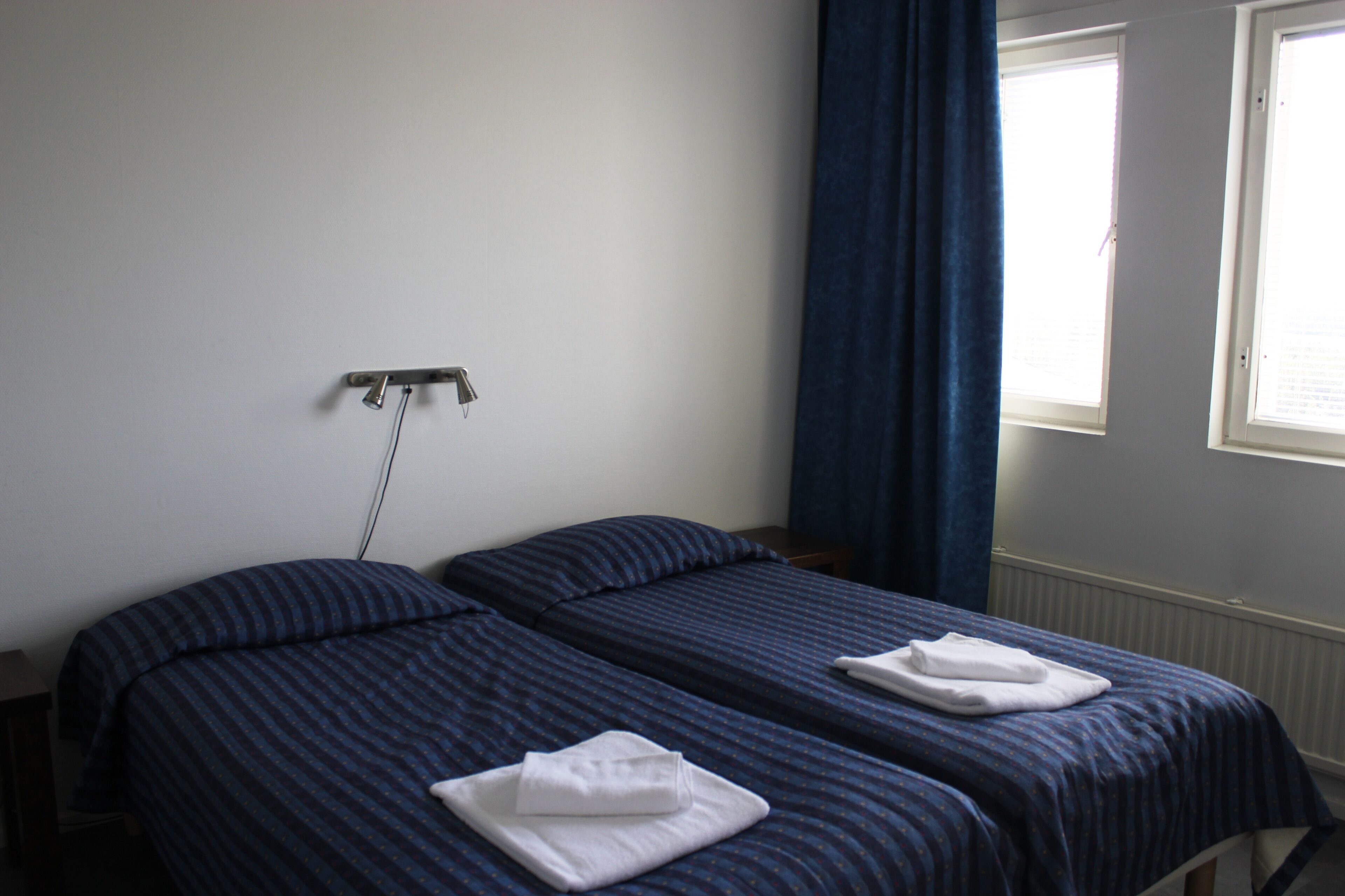 A room with two single beds, each with a folded white towel. There is a window with blinds on the right wall, and a power outlet on the left wall.
