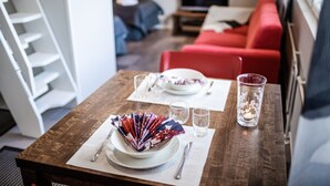 Ein Esstisch ist gedeckt mit Tellern, Besteck und Gläsern. Im Hintergrund befindet sich eine rote Couch und ein Fernseher.