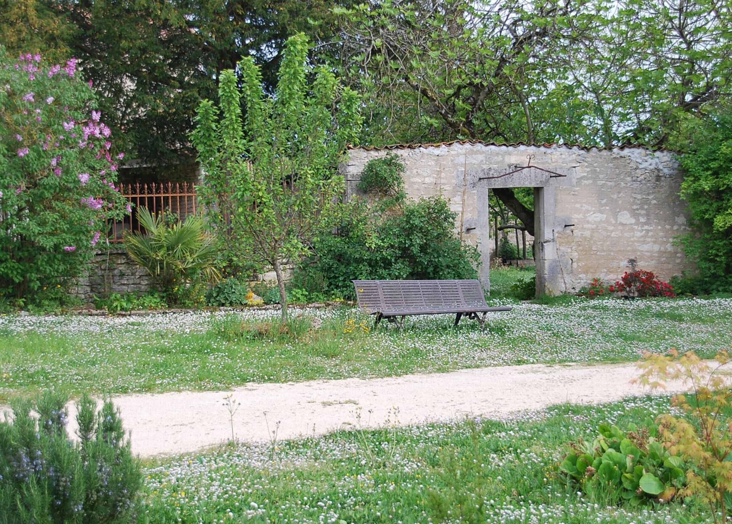 A rustic stone building with a bench in front, surrounded by lush greenery and flowering plants.