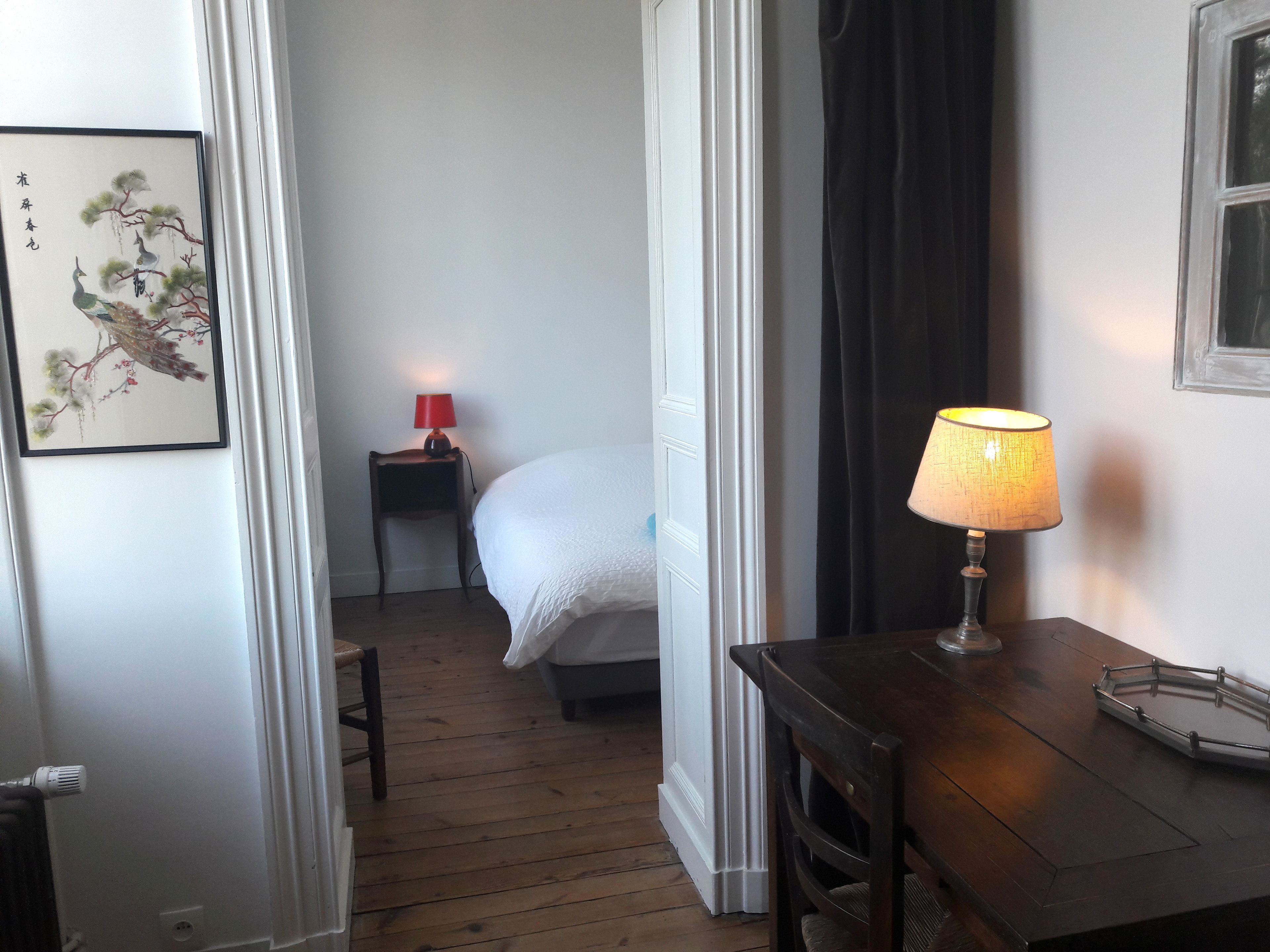 Hotellihuone, jossa on sänky, yöpöytä, jossa on punainen lamppu, puinen pöytä, jossa on lamppu, ja seinällä kehystetty taideteos.