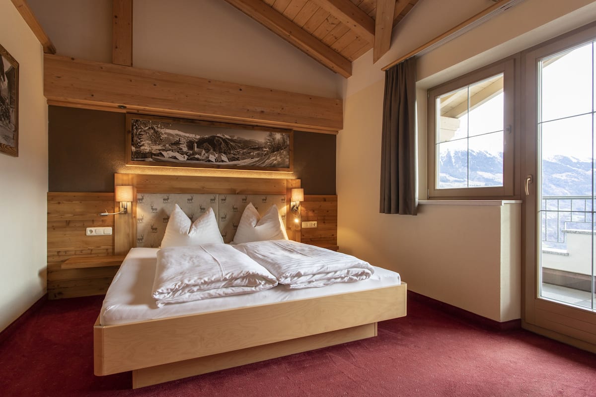 A wooden bed with white bedding in a room with a red carpet, wooden ceiling beams, a painting above the bed, and a window with a view of mountains.