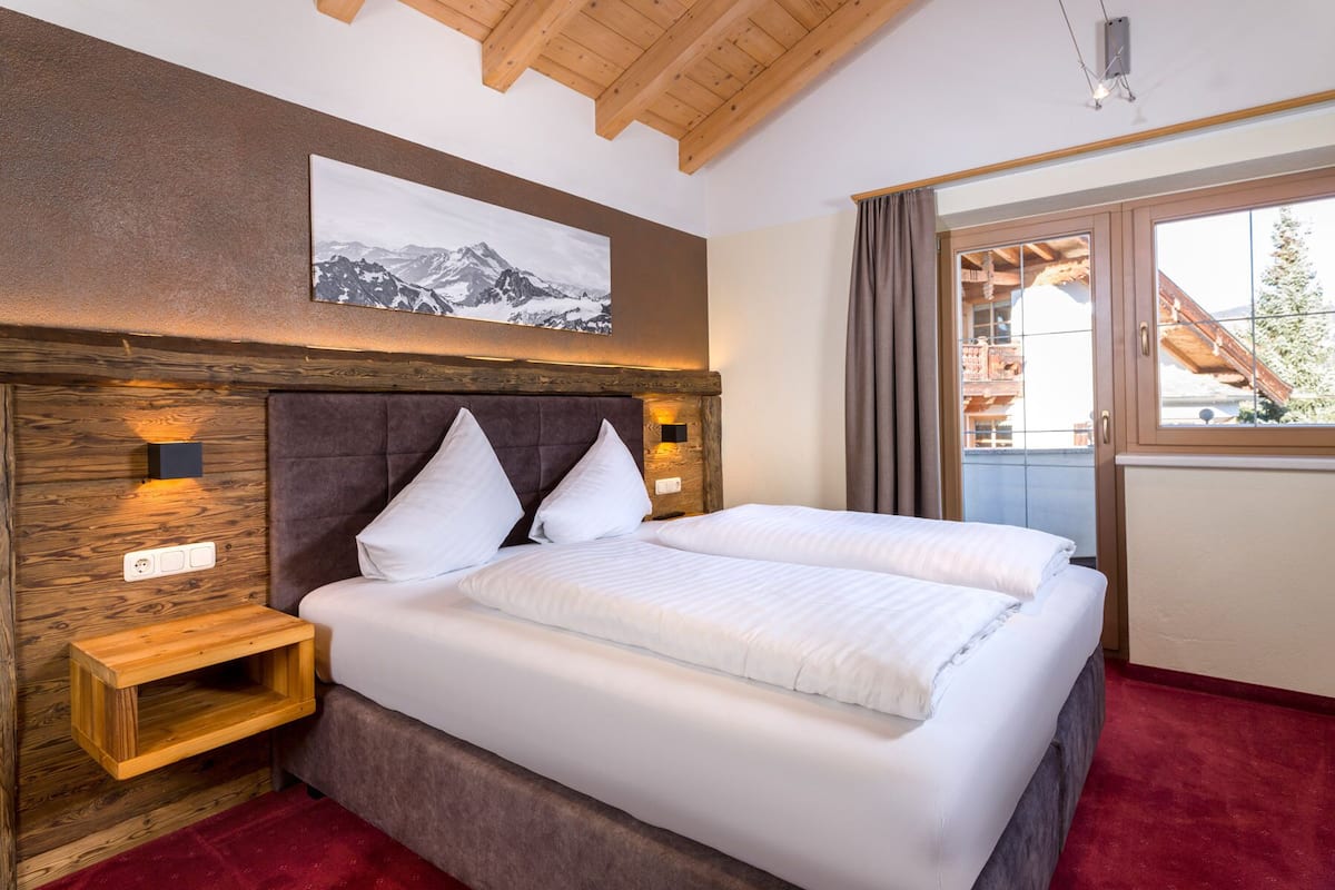 A hotel room with a wooden ceiling, a bed with white bedding, a nightstand, a wall-mounted light, and a window with curtains.