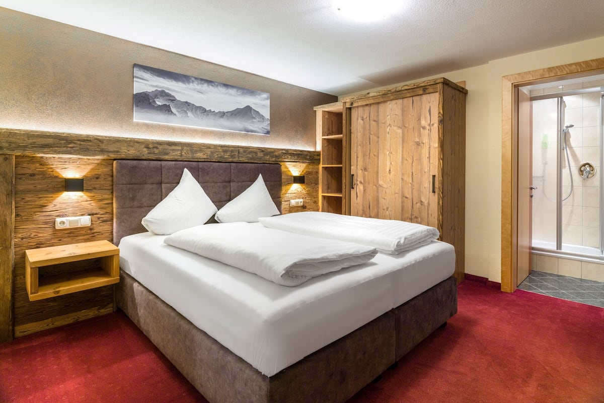 A bedroom with a large bed, wooden headboard, two bedside tables with lamps, a wardrobe, and a shower area.