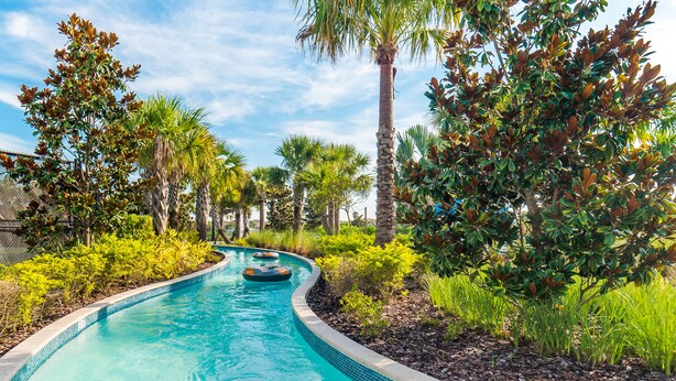 A swimming pool with a curved edge, surrounded by lush greenery and palm trees. - Fv66383 - Windsor At Westside - 5 Bed 4.5 Baths Townhome (Kissimmee)