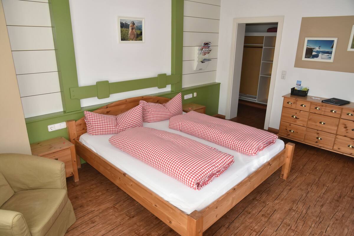 Uma cama de casal com roupa de cama xadrez, uma mesinha de cabeceira de madeira, um painel de parede verde, um penteou de madeira e uma porta que dá para um guarda-roupa.