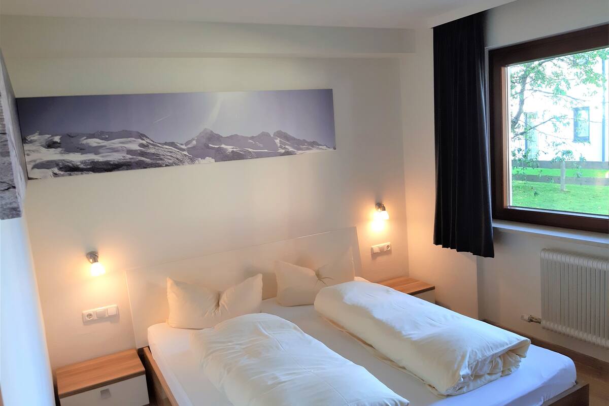 Quarto com cama, duas mesas de cabeceira, luminária na parede, janela com cortinas e pintura de paisagem de montanhas.