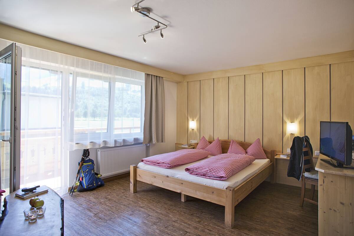 Uma cama de madeira com roupa de cama xadrez, uma escrivaninha com um computador e uma janela com cortinas.