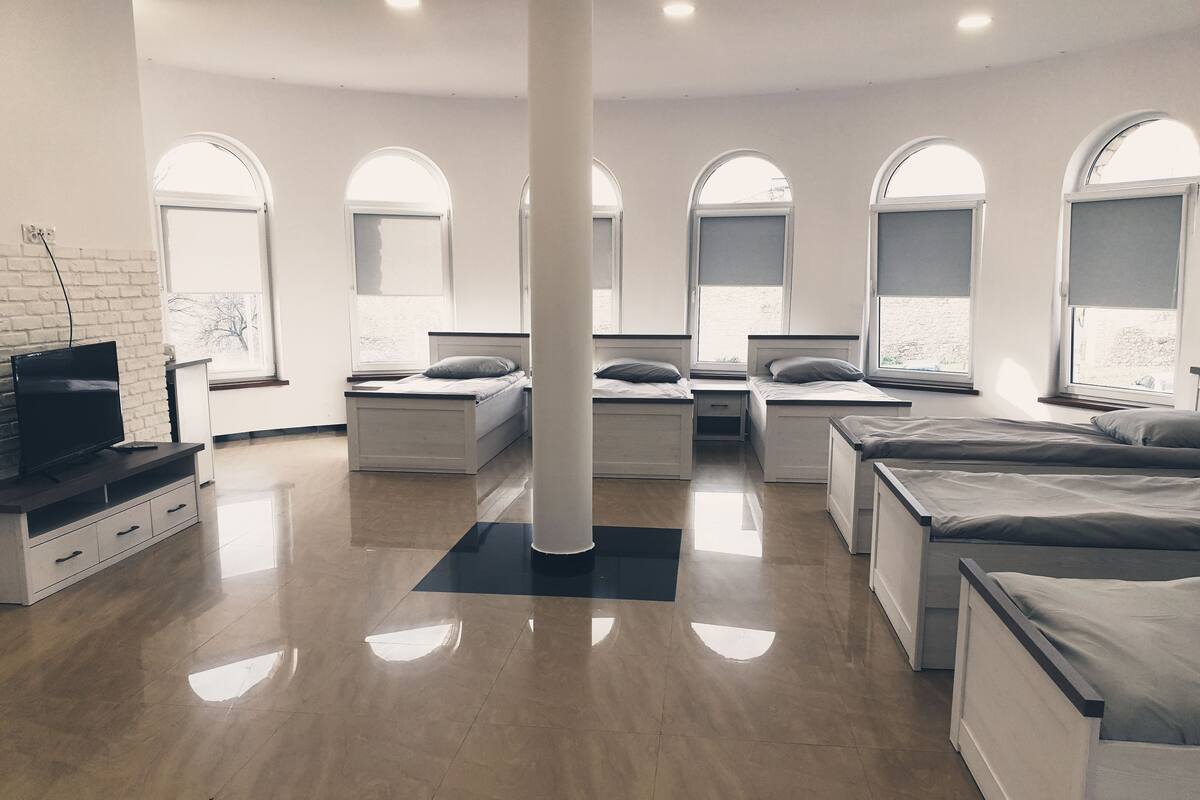 A modern, well-lit room with a large central column, multiple beds, a TV on a wooden console, and large arched windows.