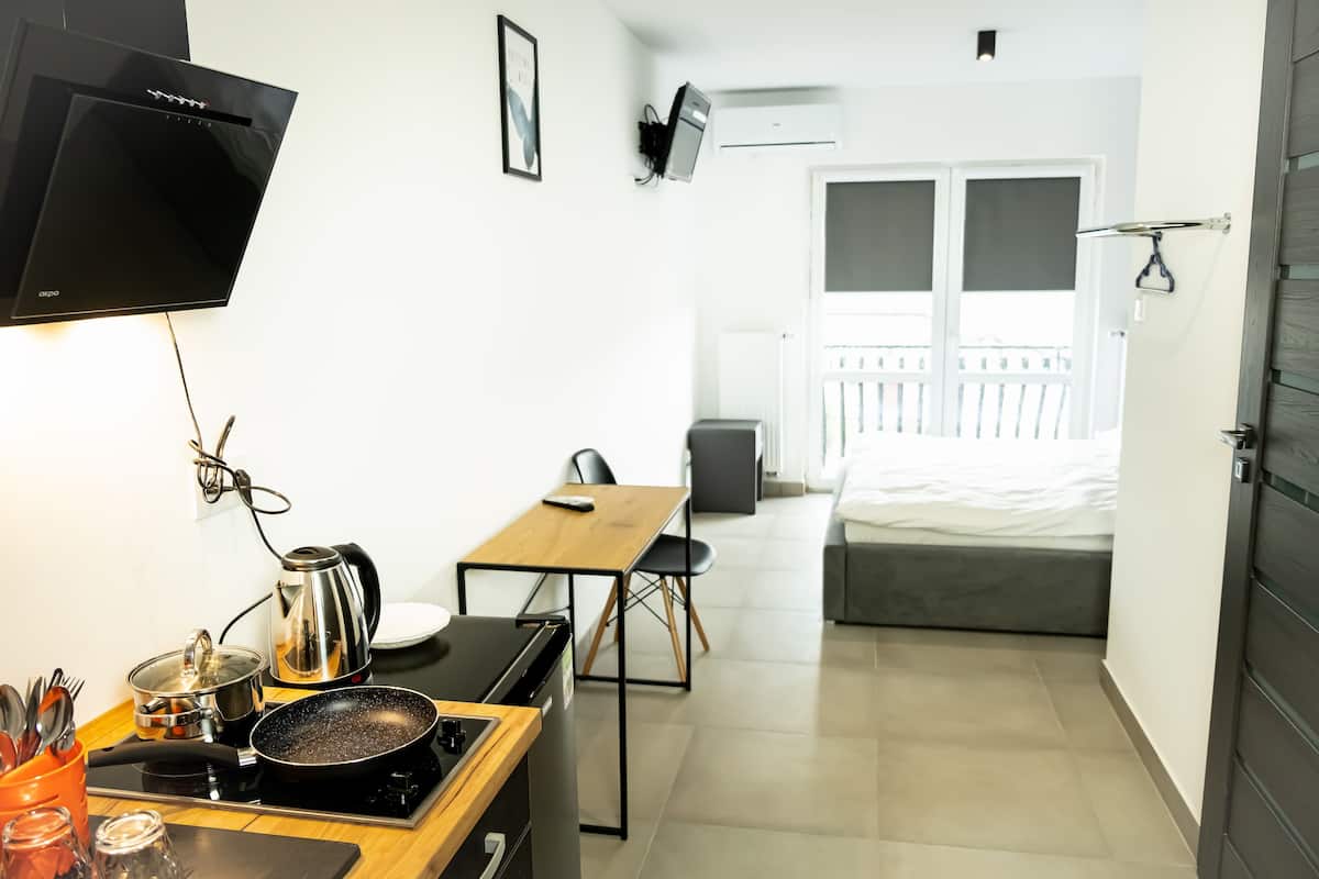 A compact kitchen with a stove, kettle, and sink, a dining area with a table and chairs, and a bed with a balcony view.
