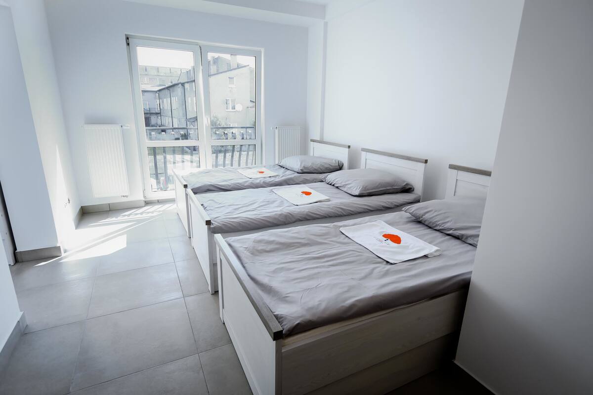 A modern, minimalist bedroom with three beds aligned against a wall, a large window providing natural light, and a view of the cityscape.