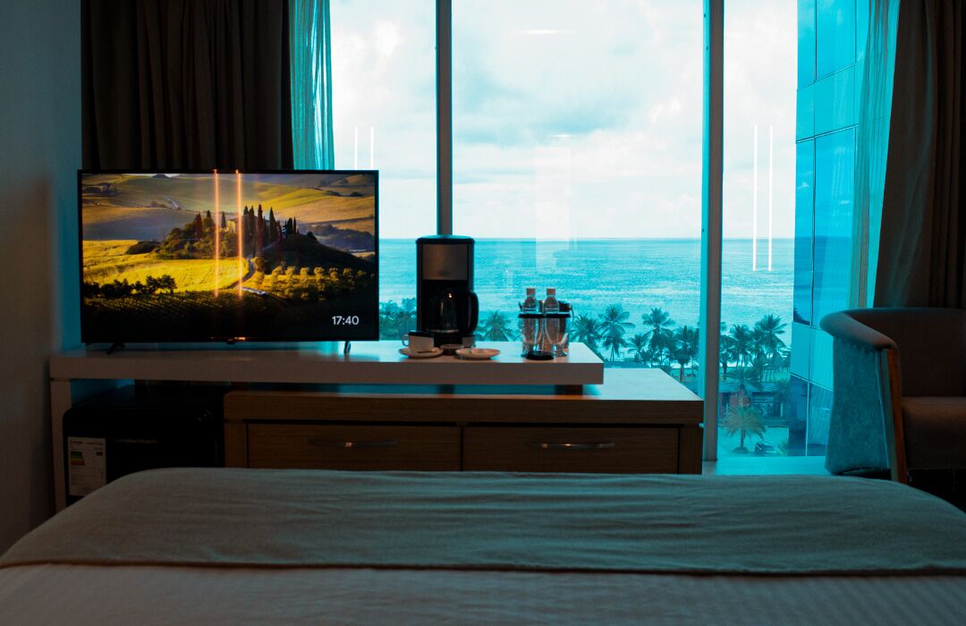 A hotel room with a large TV on a wooden console, a coffee maker, and a view of the ocean through large windows.