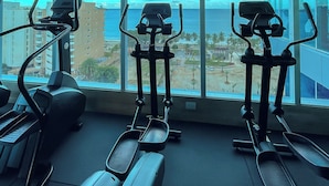Ein Fitnessraum mit Laufbändern und Crosstrainern, große Fenster mit Blick auf Stadt und Meer.