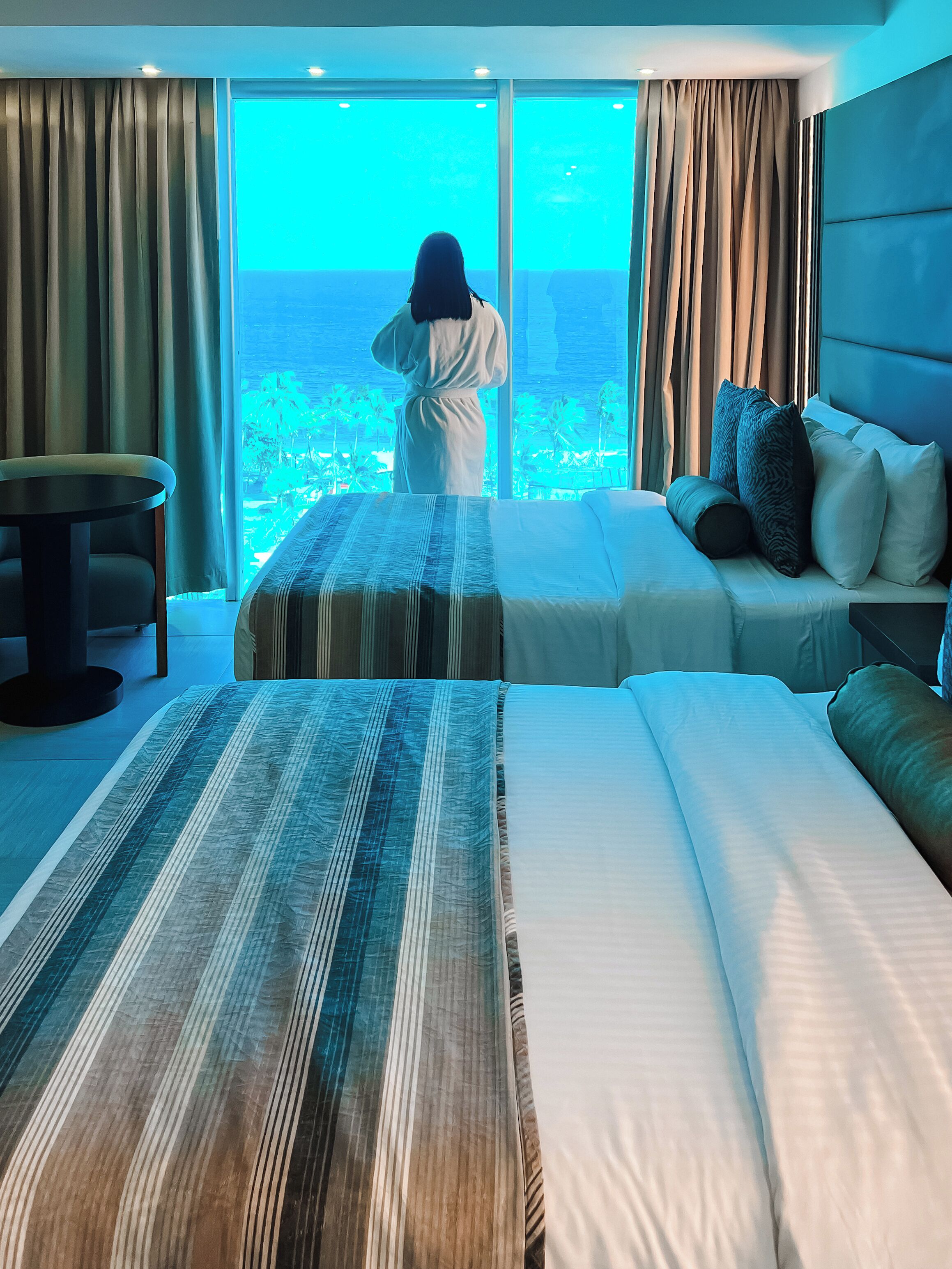 A hotel room with a large window, a bed with striped bedding, a sofa with pillows, and a small table.